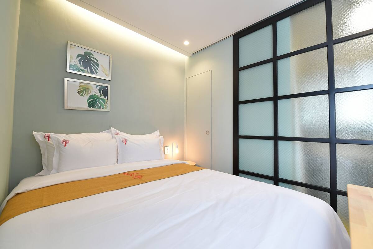 Un lit bien fait, avec du linge de lit blanc et un plaid jaune, une lampe fixée au mur et deux œuvres d’art encadrées représentant des feuilles tropicales.