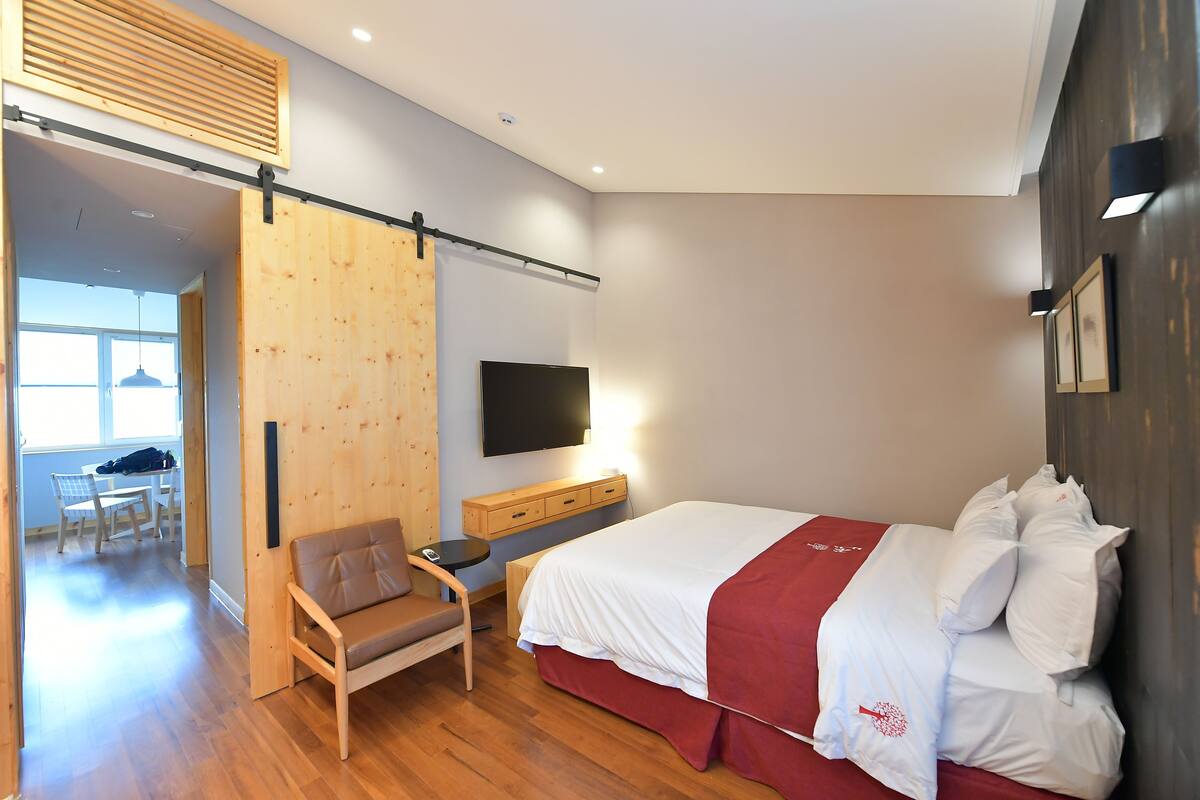 Une chambre d’hôtel moderne avec un plancher en bois, un lit avec une literie blanche et un détail rouge, un bureau avec une chaise, un téléviseur à écran plat fixé au mur et une cloison coulissante en bois.