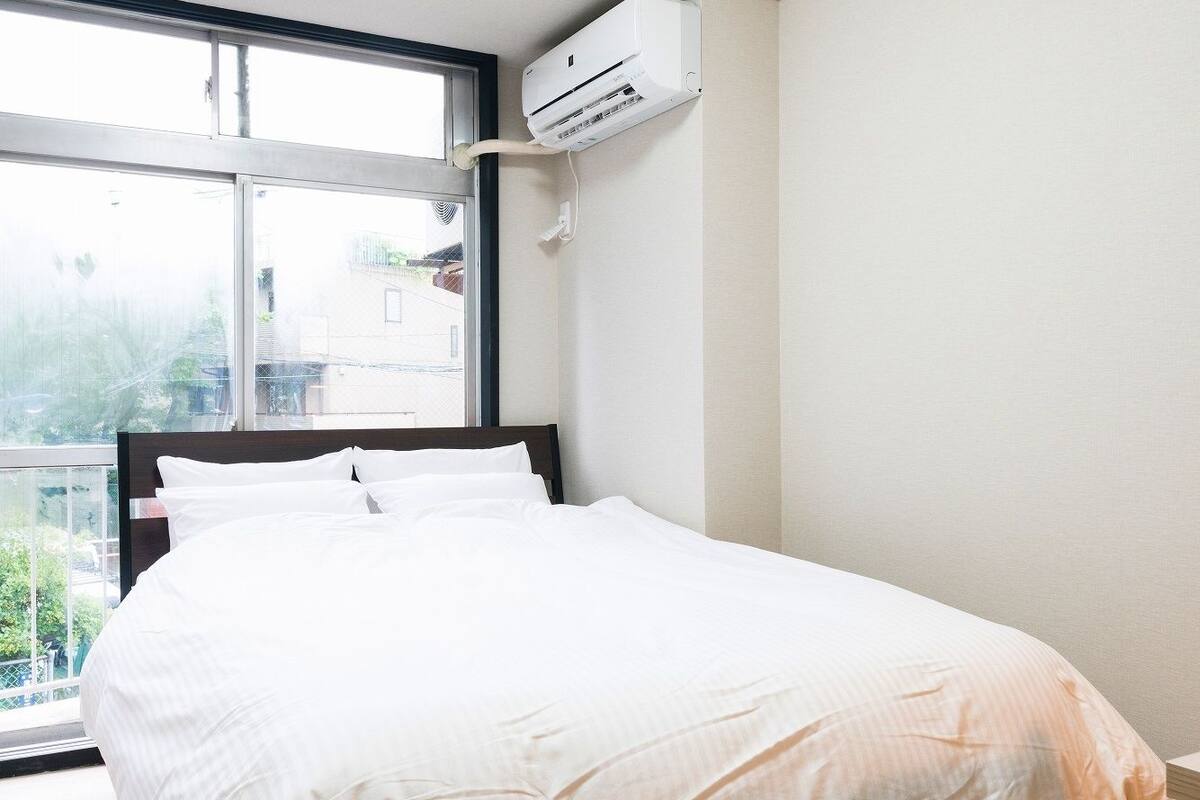 Un dormitorio con cama, piso de madera, paredes blancas, una ventana y un equipo de aire acondicionado.
