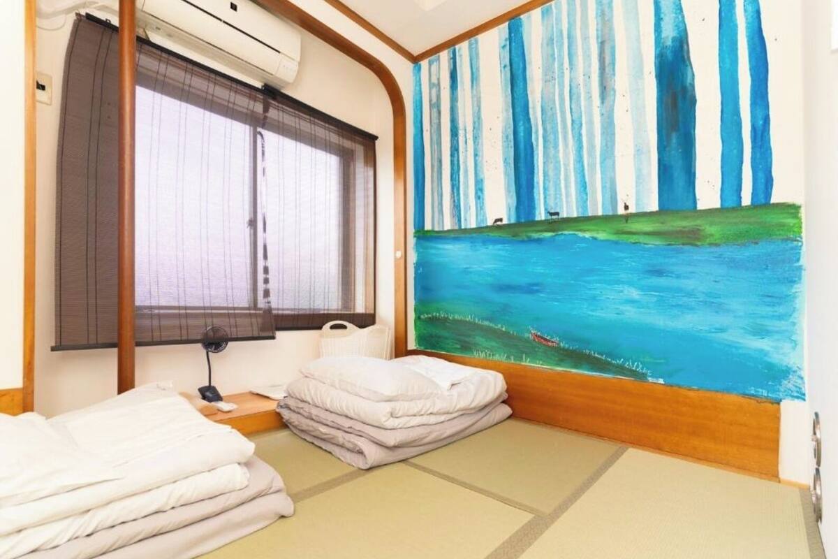 Una habitación con cama, ventana con persianas, un cuadro de un río y un ventilador.