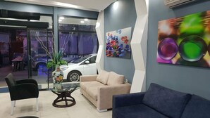 Un área de recepción moderna con un sofá beige, un sofá azul y una mesa de centro de vidrio. Se ve un coche a través de la puerta de cristal, y un cuadro abstracto colorido en la pared.