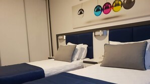 Habitación de hotel con dos camas, cada una con cubrecama azul y ropa de cama blanca. Hay dos mesitas de noche con lámparas, y un aplique en la pared sobre las camas.