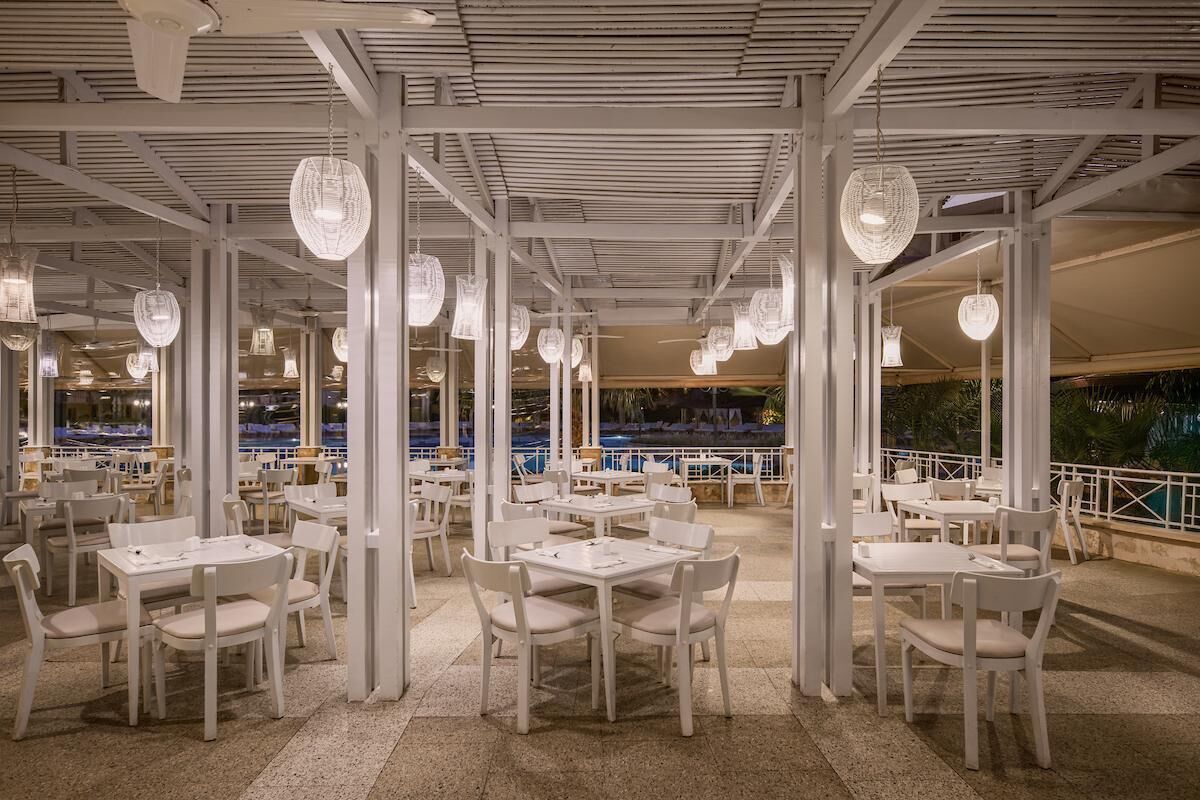 Een terras met witte tafels en stoelen, hangende lampen en uitzicht op een zwembad.