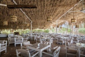 An outdoor dining area with white tables and chairs, a thatched roof, and hanging light fixtures.