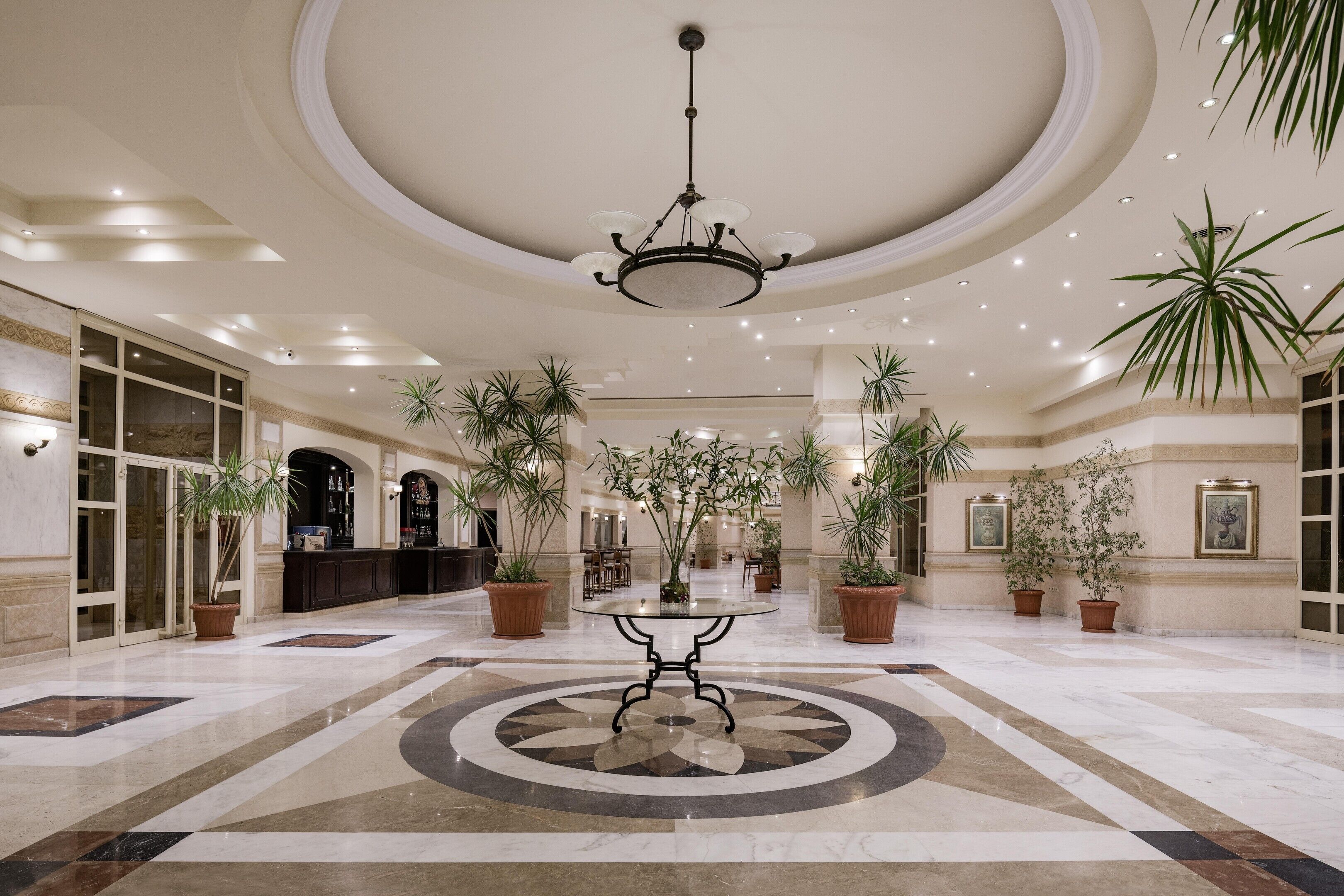 A spacious lobby with a central round table, multiple potted plants, and a chandelier.