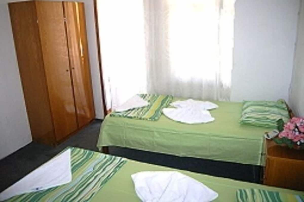 A room with two beds, each with green bedding and white pillows. There is a wooden wardrobe and a small table with a vase of flowers.
