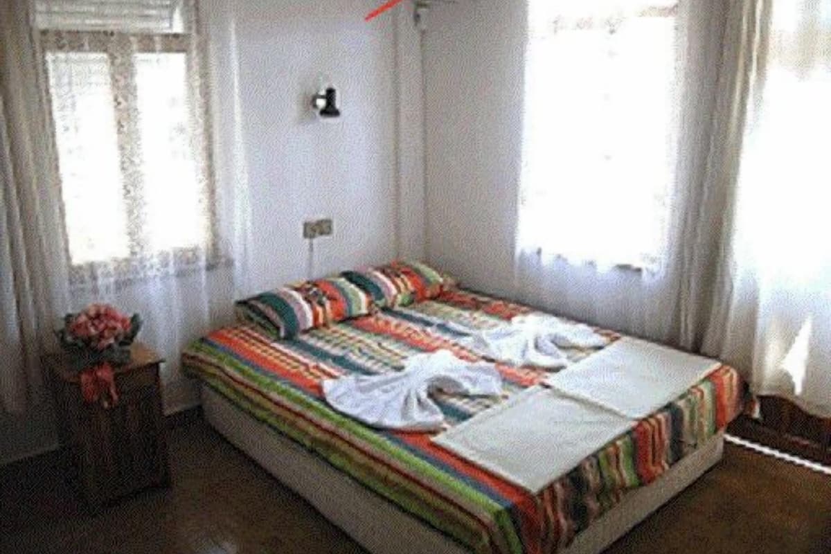 A single bed with a colorful striped bedspread, a bedside table with a vase of flowers, and a window with sheer curtains.