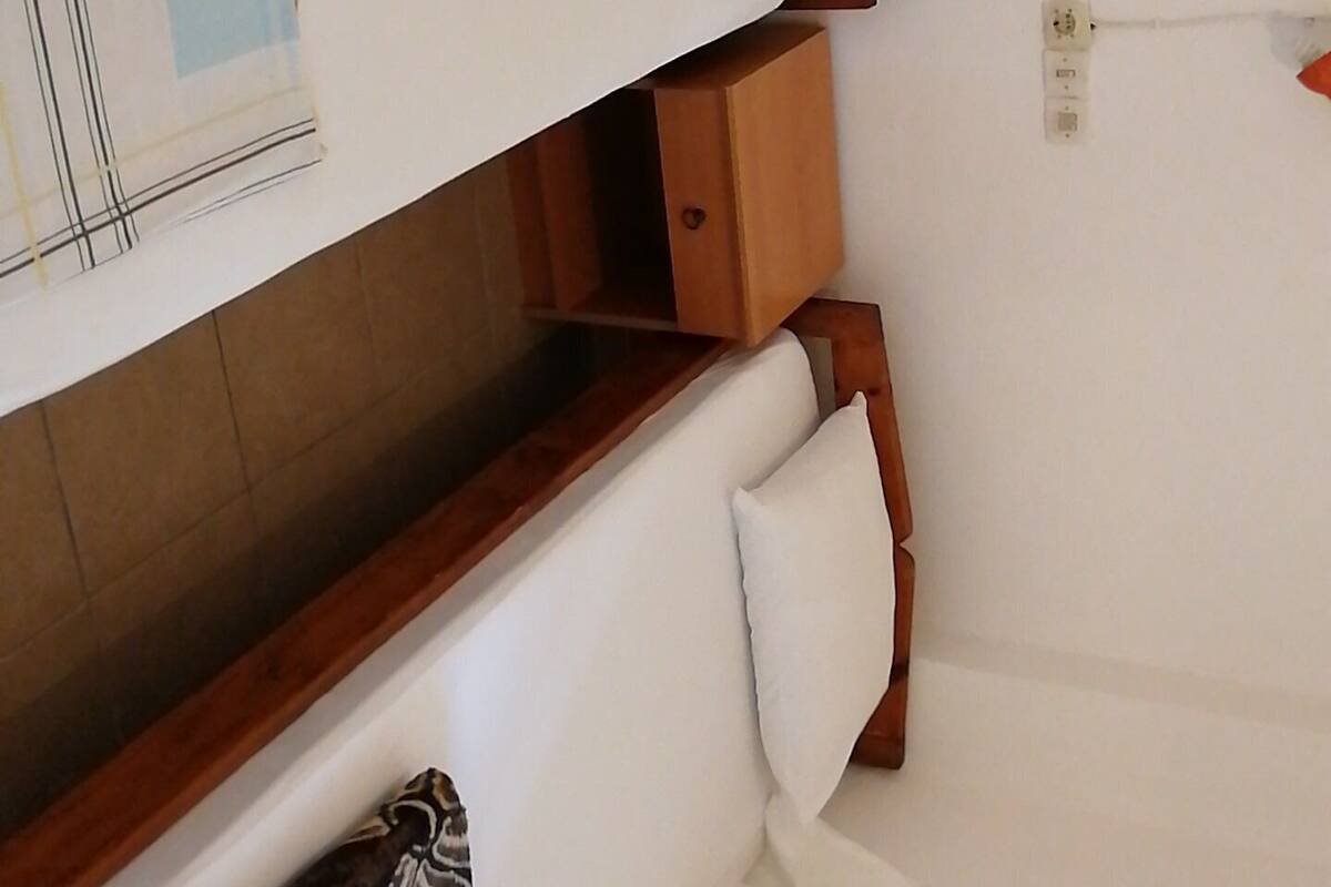 A compact, single-bed room with a wooden headboard, a small wooden cabinet, and a wall-mounted electrical outlet.