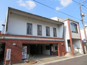 A two-story building with a white facade and red brick base. There is a sign on the ground floor indicating a business, and a no parking sign is visible on the right side. - Tsuyama Grand Hotel (Tsuyama)