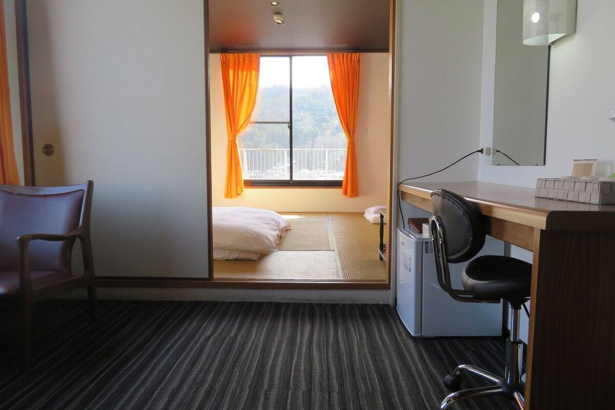 Une chambre d’hôtel compacte comprenant un lit, un bureau avec une chaise, un petit réfrigérateur et une fenêtre avec des rideaux orange.