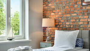 A hotel room with a brick wall, a bed with white bedding, a nightstand with a lamp, and a window with a sailboat model. - Oxford Family apartment in a quiet Sopot townhouse by Lion Apartments (Sopot)