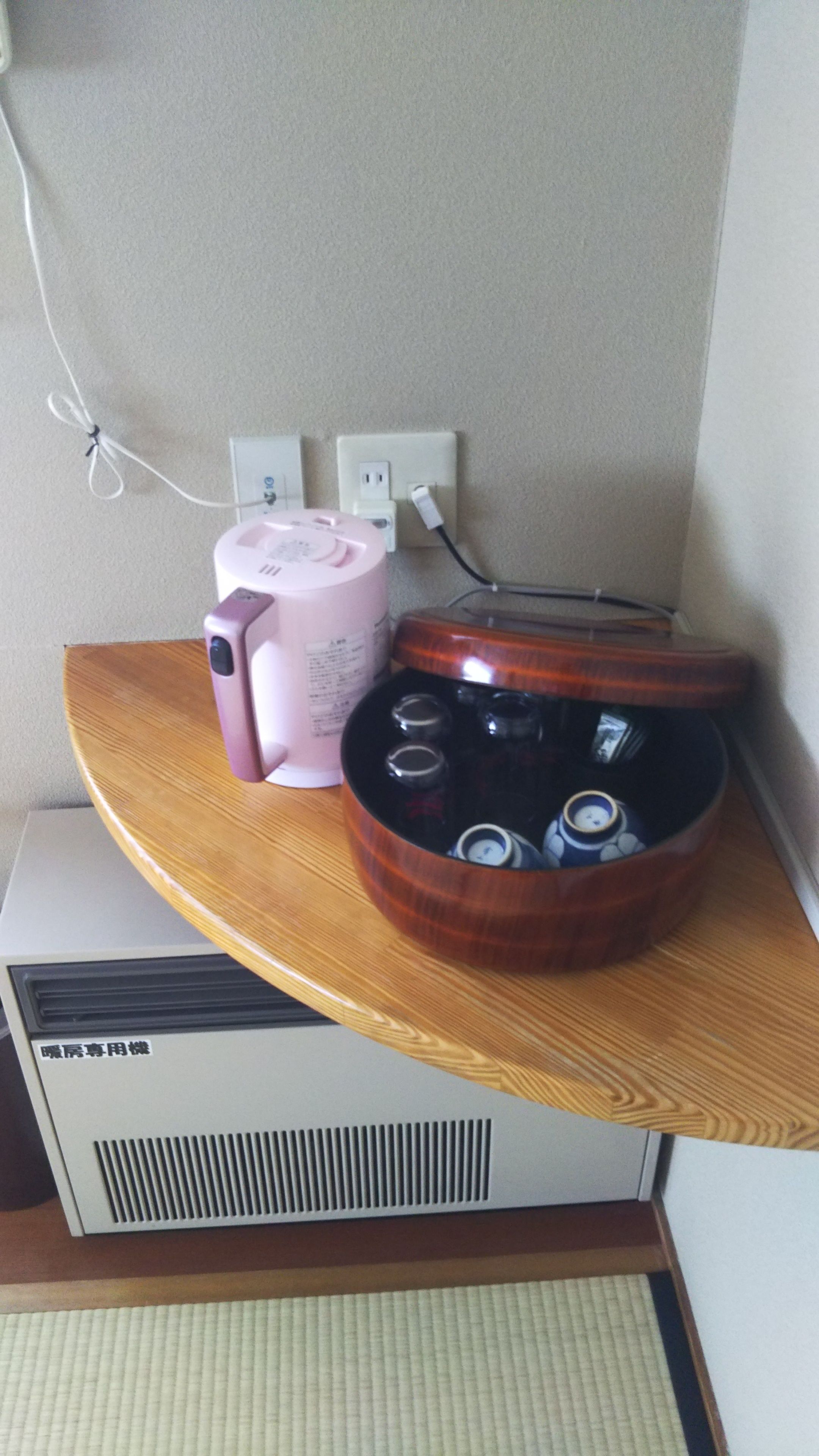 A corner of a room with a wooden shelf holding a pink device, a black container with multiple lids, and a white appliance with ventilation grilles.