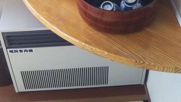 A corner of a room with a wooden shelf holding a pink device, a black container with multiple lids, and a white appliance with ventilation grilles.
