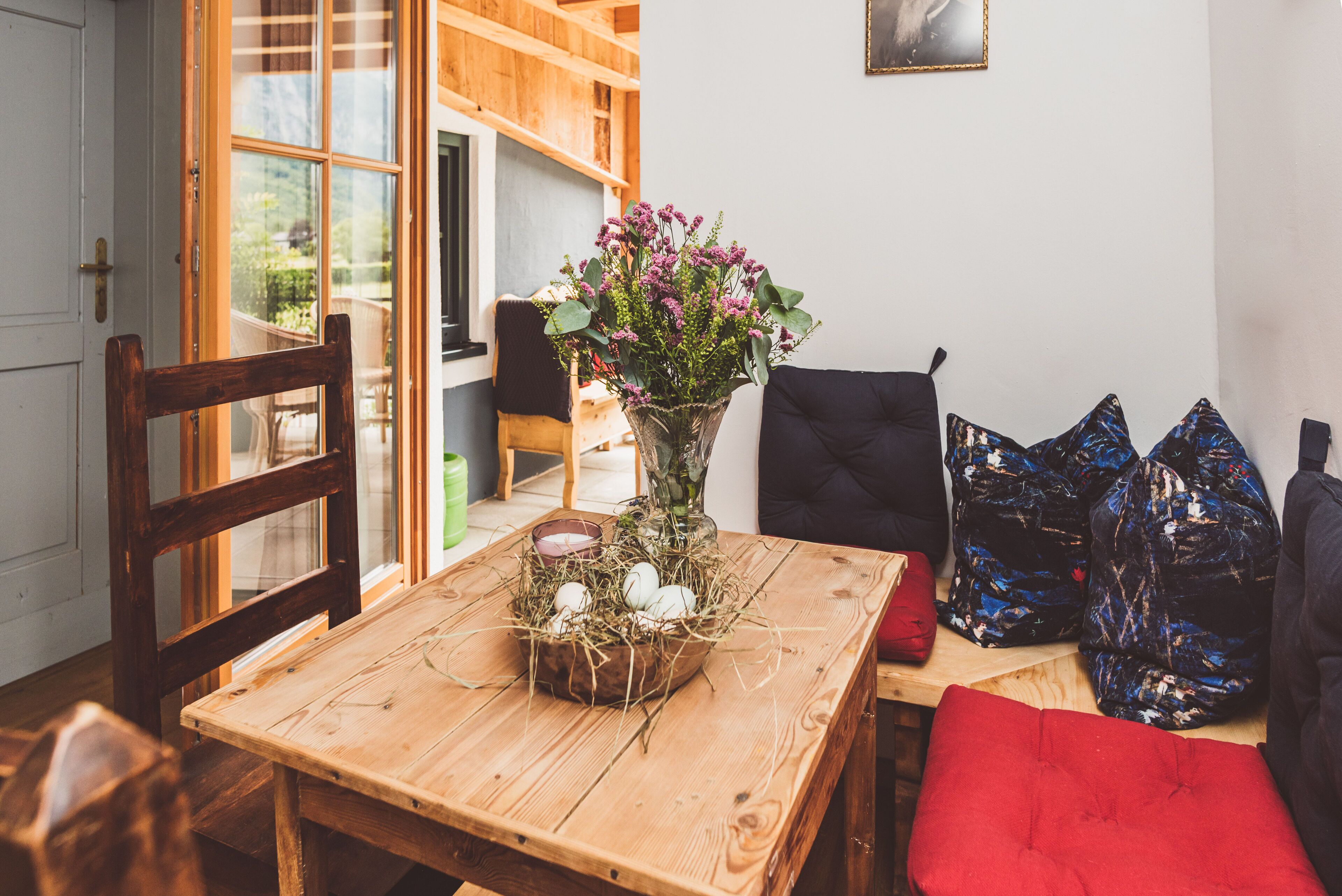 Une table de salle à manger en bois, ornée d’un vase de fleurs, d’un panier rempli d’œufs et d’un coussin rouge.