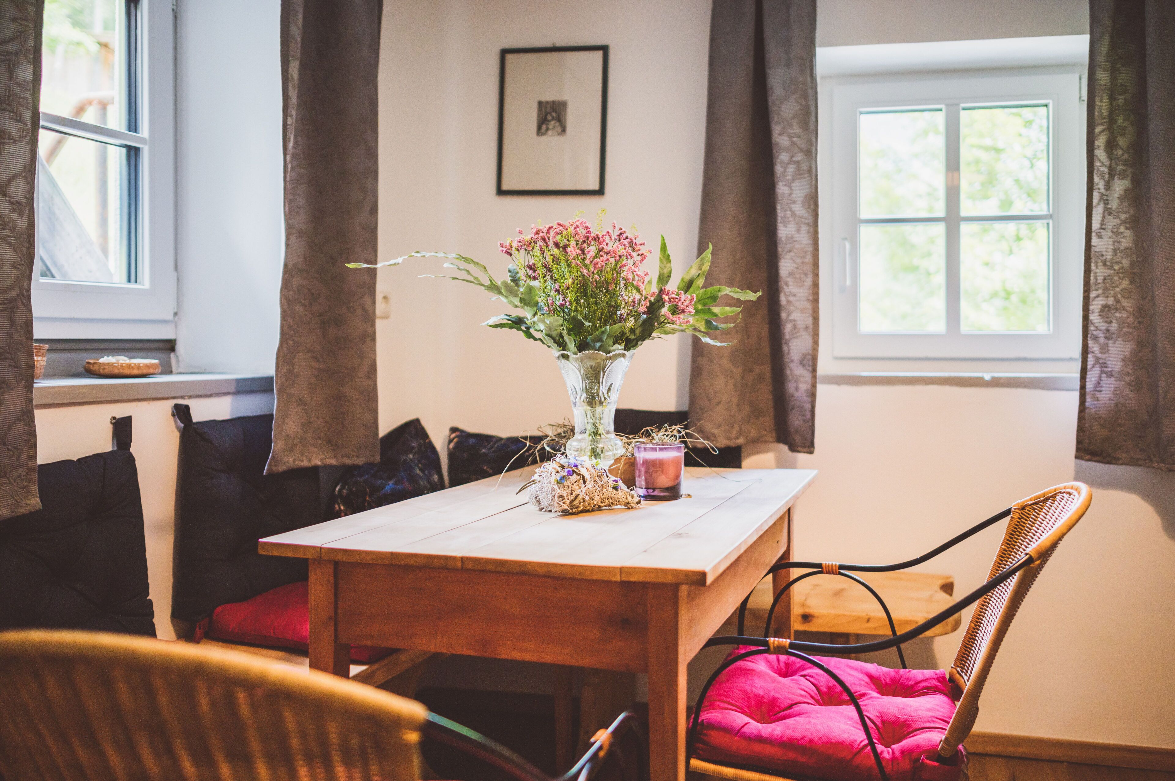 A wooden dining table with a vase of flowers, a candle, and a basket. There are chairs with cushions around the table.