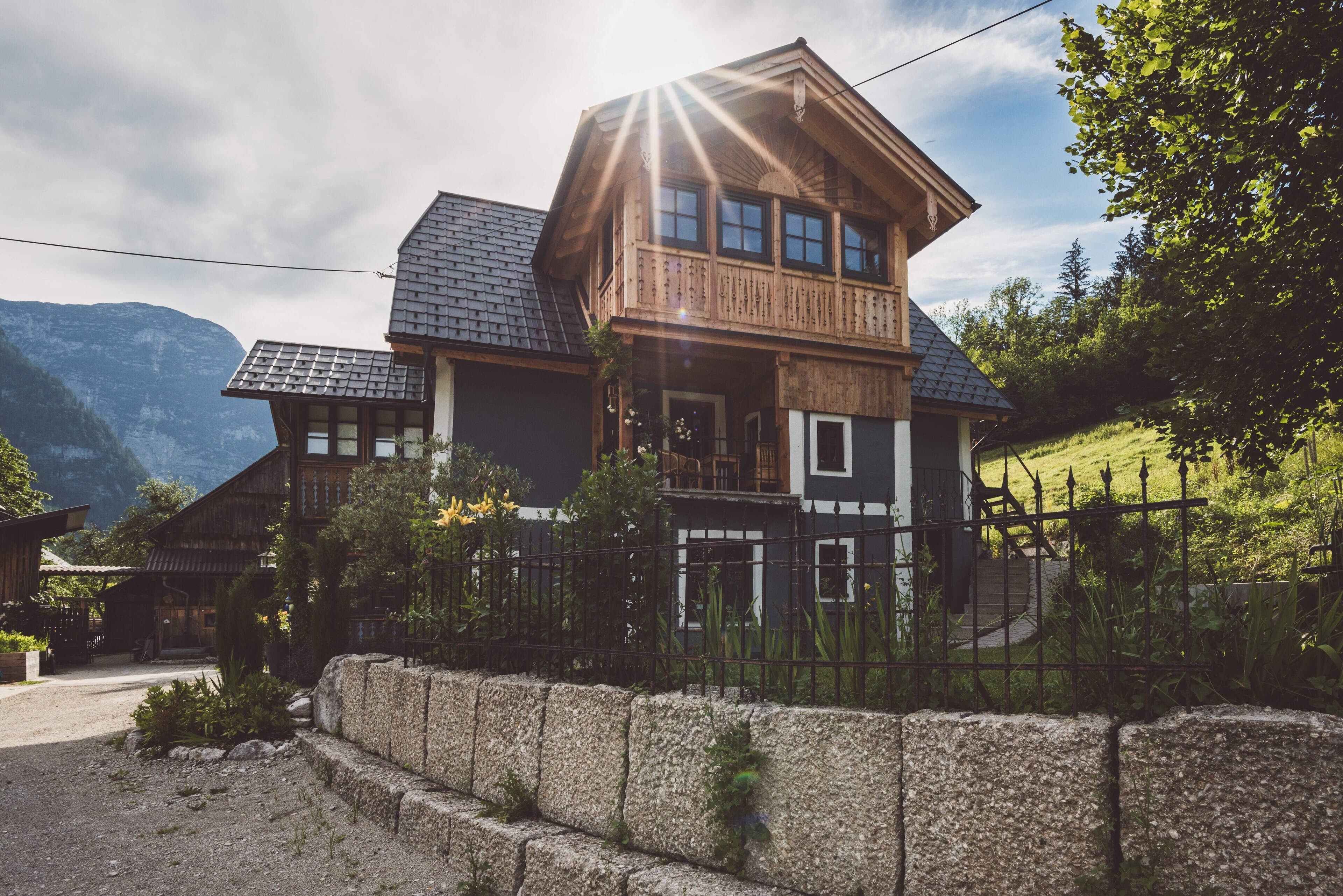 A wooden house with a balcony, surrounded by a stone wall and a metal fence, set against a mountainous backdrop.