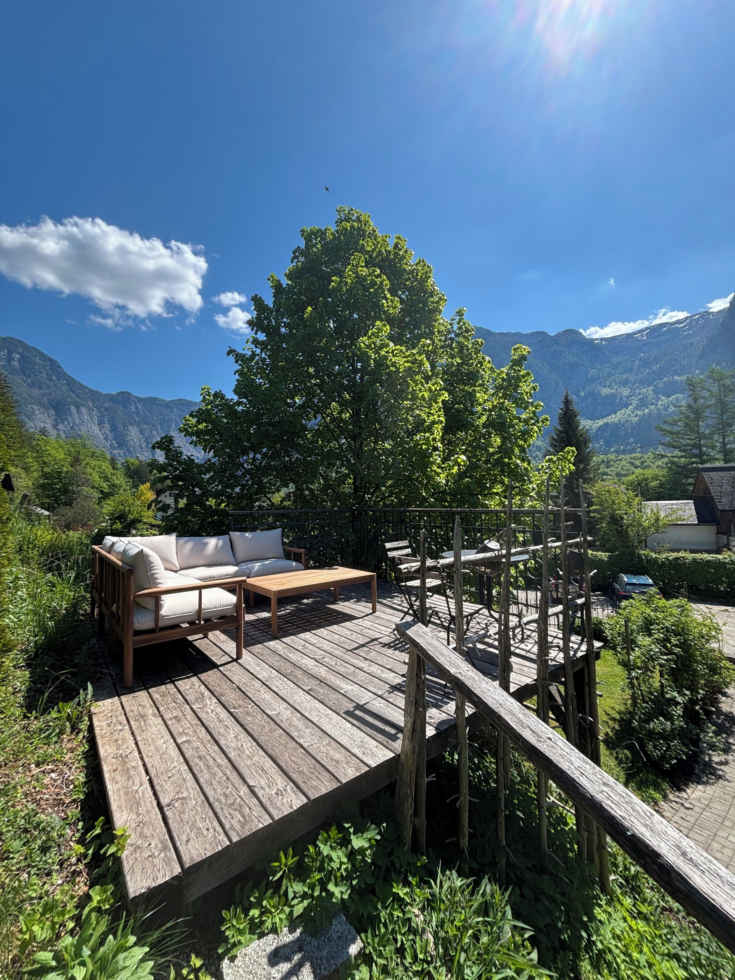 Eine Holzterrasse mit einer Couch und einem Tisch, umgeben von Grünpflanzen und Bergen im Hintergrund.