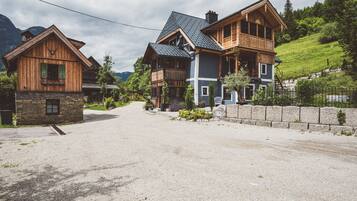 A two-story wooden house with a stone foundation, a gravel driveway, and a hillside with greenery.