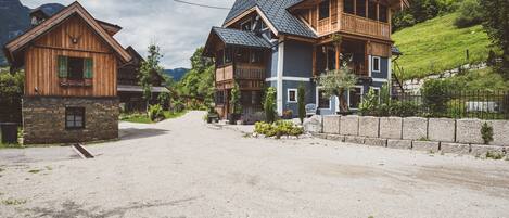 A two-story wooden house with a stone foundation, a gravel driveway, and a hillside with greenery.