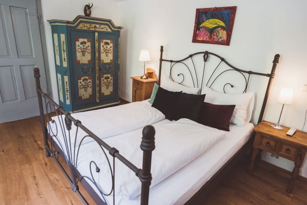 Un dormitorio con estructura de cama metálica, armario de madera, mesita de noche con lámpara y un cuadro colorido en la pared.