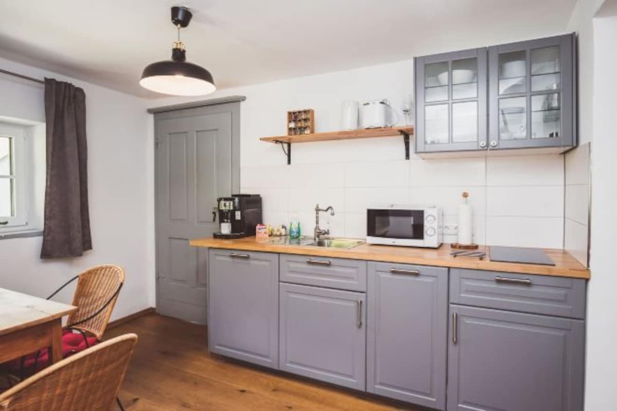 Cocina compacta con gabinetes grises, encimera de madera, microondas, cafetera y una lámpara colgante.
