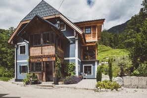 Una casa de madera de dos pisos con un balcón, rodeada de follaje y colinas.