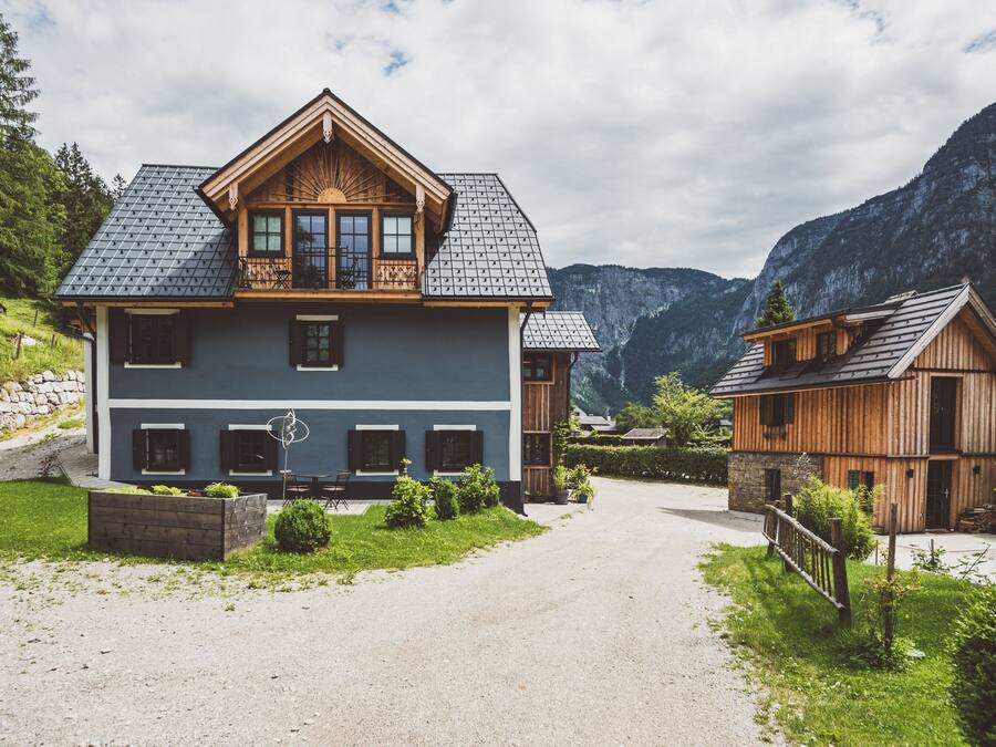 Une maison en bois à deux étages, dotée d’un balcon, d’une allée en gravier et surplombant un paysage montagneux.