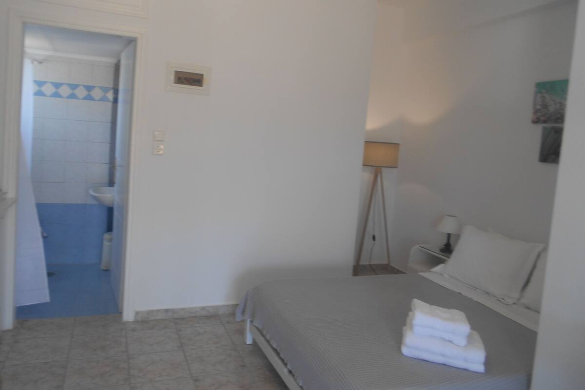A bedroom with a bed, a bedside table, a lamp, and a bathroom visible through an open door.