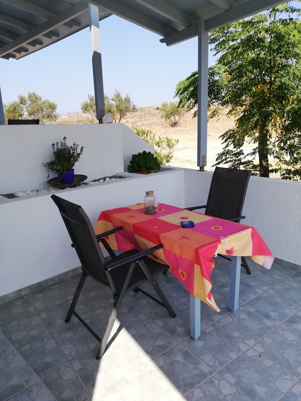 An outdoor patio with a table covered in a colorful tablecloth, two chairs, and potted plants.