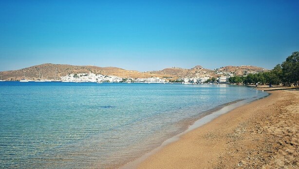 A coastal beach with clear blue water, sandy shore, and white buildings in the background. - Elea Milos (Milos)