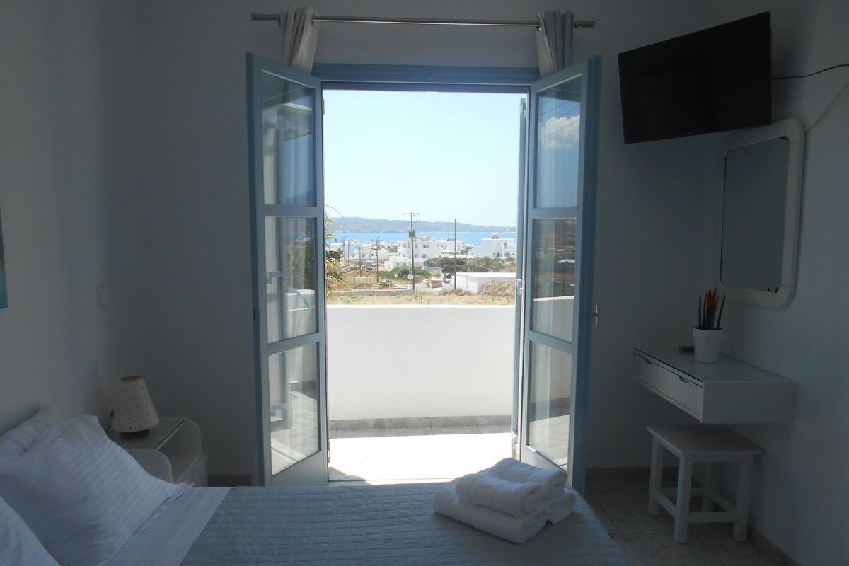 A bedroom with a bed, a bedside table, a nightstand, a window with an open door, and a view of the outside.