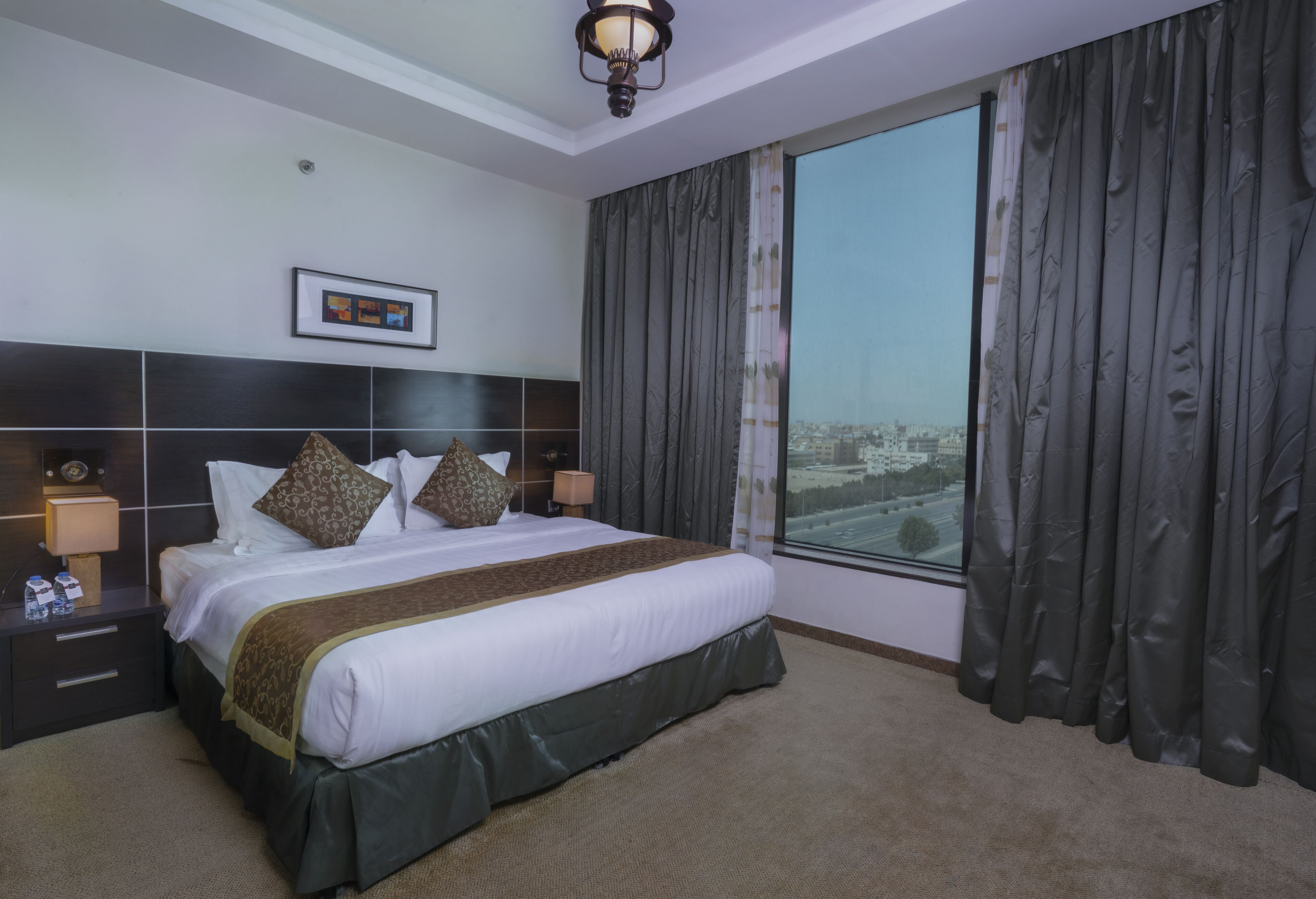 A hotel room with a large bed, dark curtains, a city view, and a framed picture on the wall.