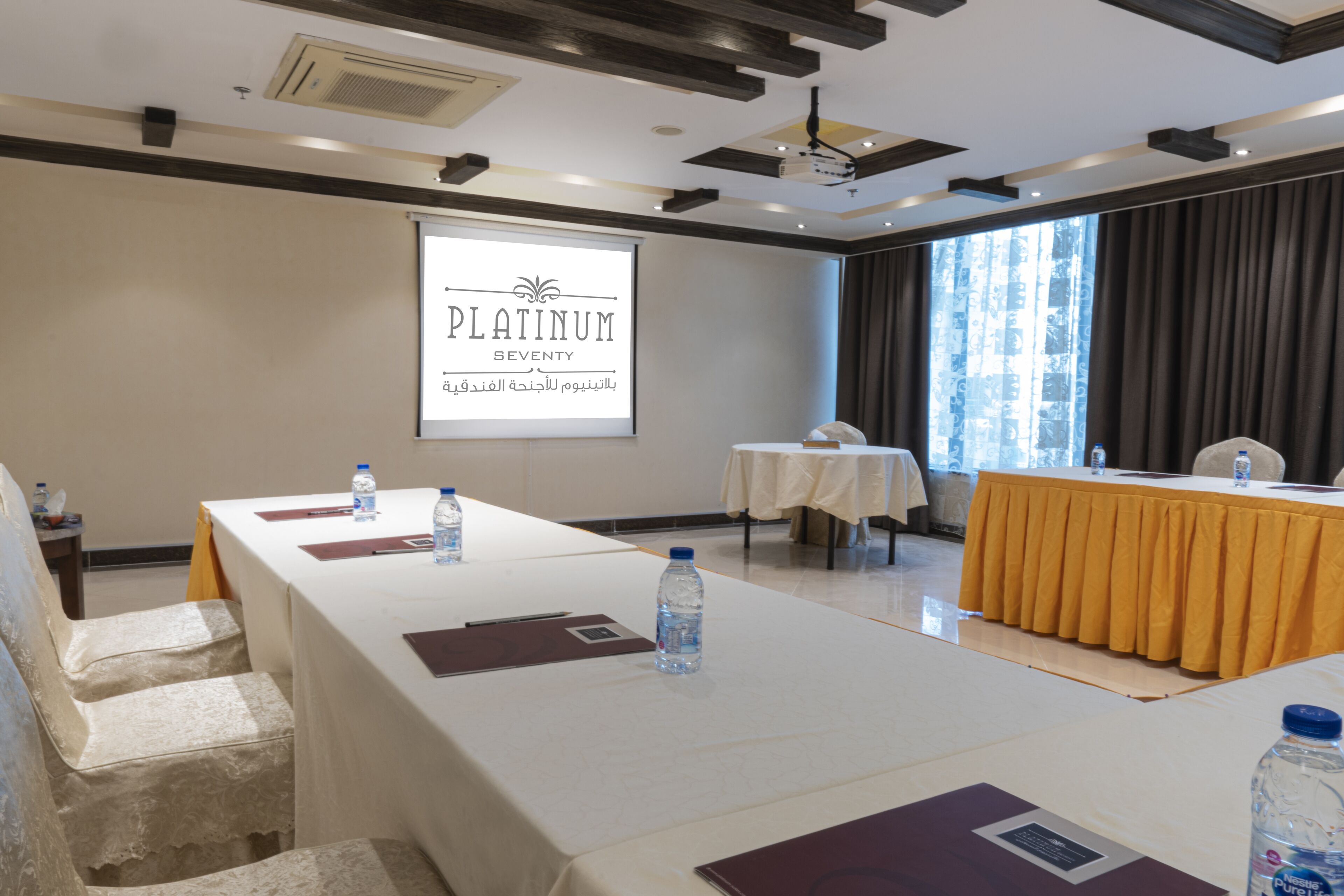 A conference room with a long table, water bottles, and a projector screen displaying "PLATINUM SEVENTY".