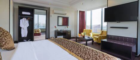 A hotel room with a large bed, a desk, a chair, a sofa, a TV, and a view of the city.