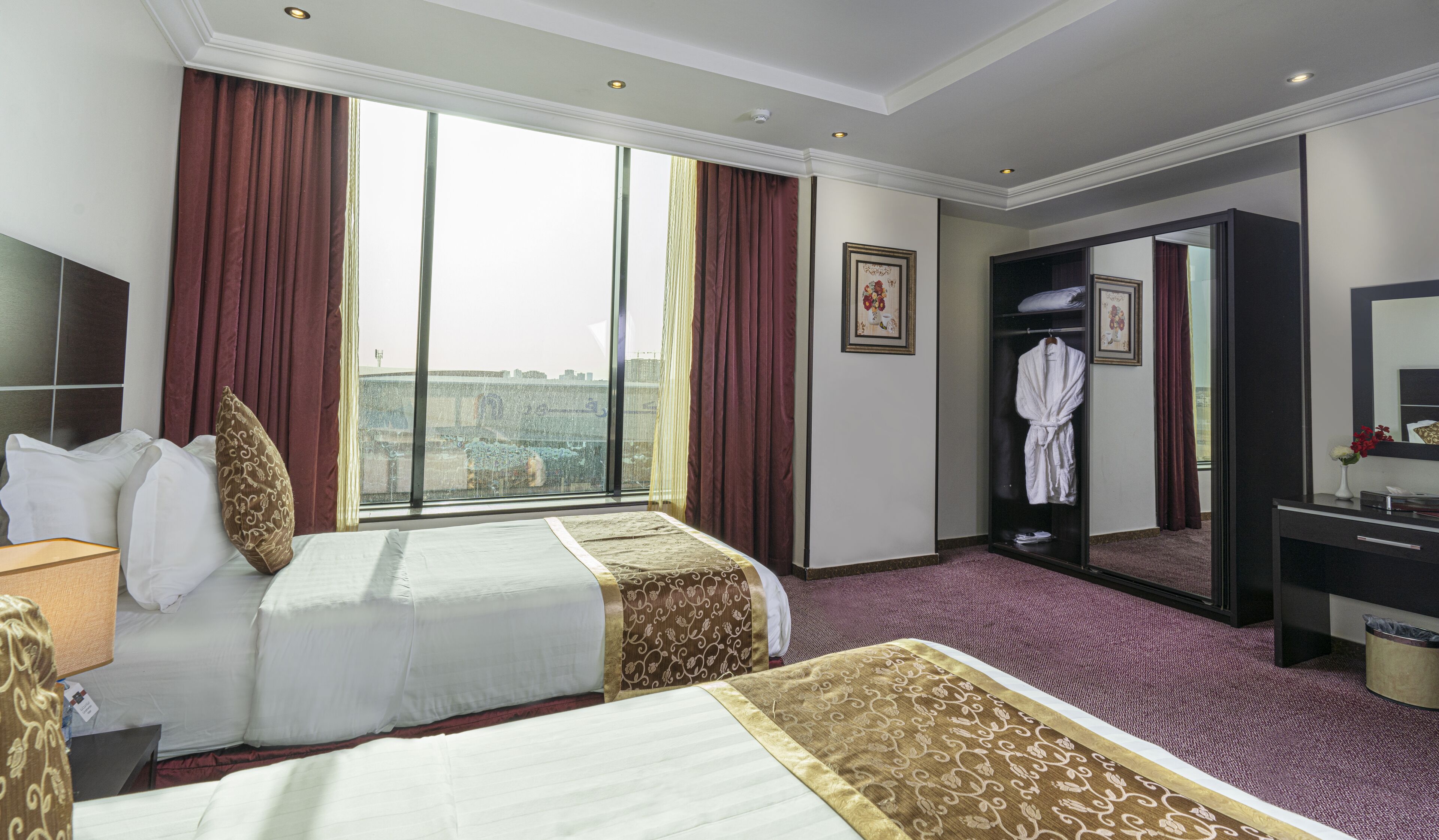 Habitación de hotel con dos camas, un ventanal con cortinas, una bata colgada en la puerta y un escritorio con espejo.