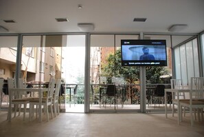 A modern indoor space with a large TV screen displaying a program, glass doors leading to an outdoor area with chairs and tables, and a view of buildings and trees outside. - Bursa Park Apart Hotel (Bursa)