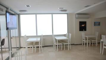 Una sala de conferencias con mesas y sillas blancas, ventanas amplias y un equipo de aire acondicionado en la pared.