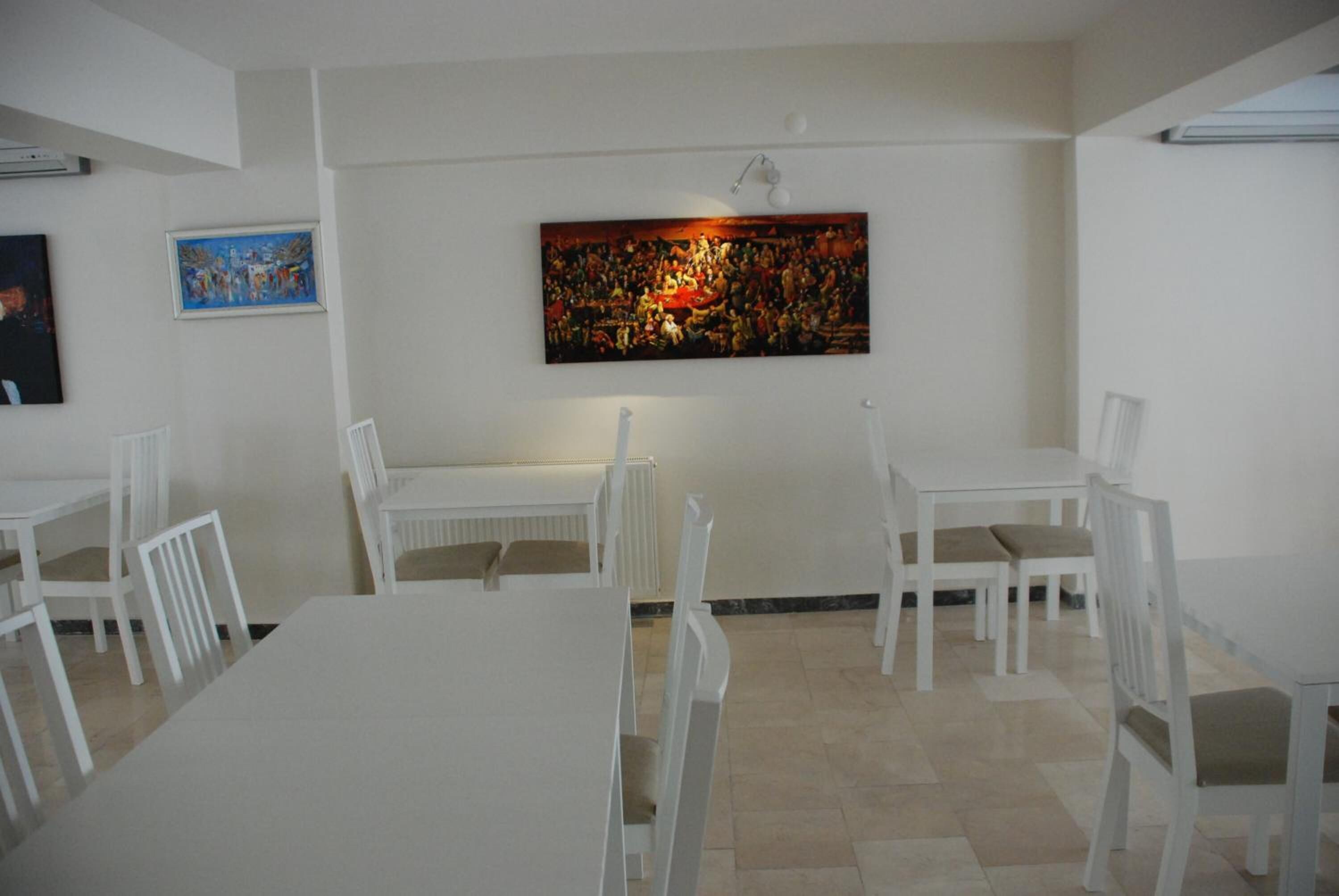 A dining area with white tables and chairs, a large painting on the wall, and a ceiling light fixture.