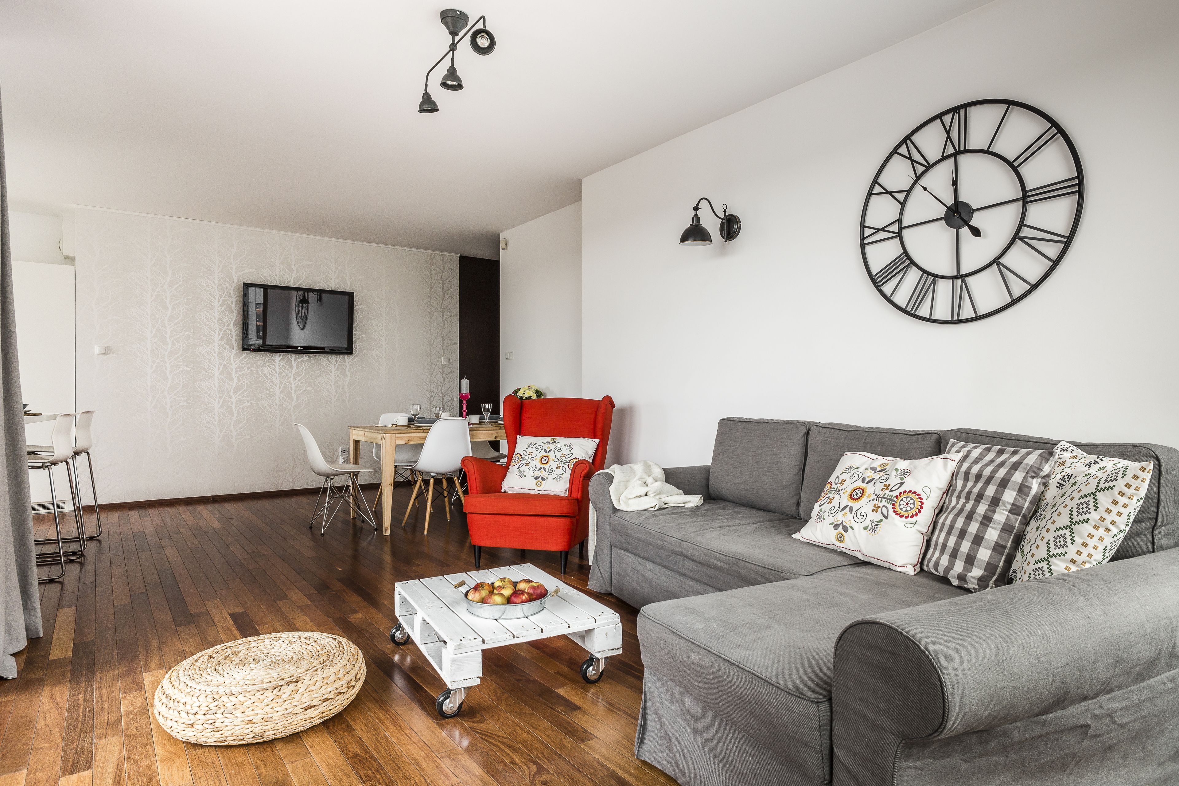 Een woonkamer met een grijze bank, een rode fauteuil, een ronde wandklok en een aan de muur bevestigde televisie.