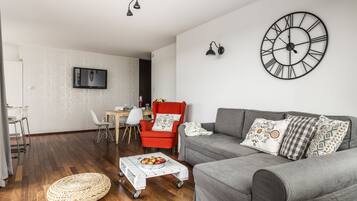 Een woonkamer met een grijze bank, een rode fauteuil, een ronde wandklok en een aan de muur bevestigde televisie.