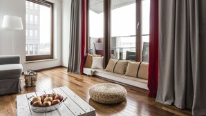 Un salon moderne avec un canapé, une table basse en bois sur laquelle sont posées des pommes, un pouf en osier et une grande fenêtre avec des rideaux.