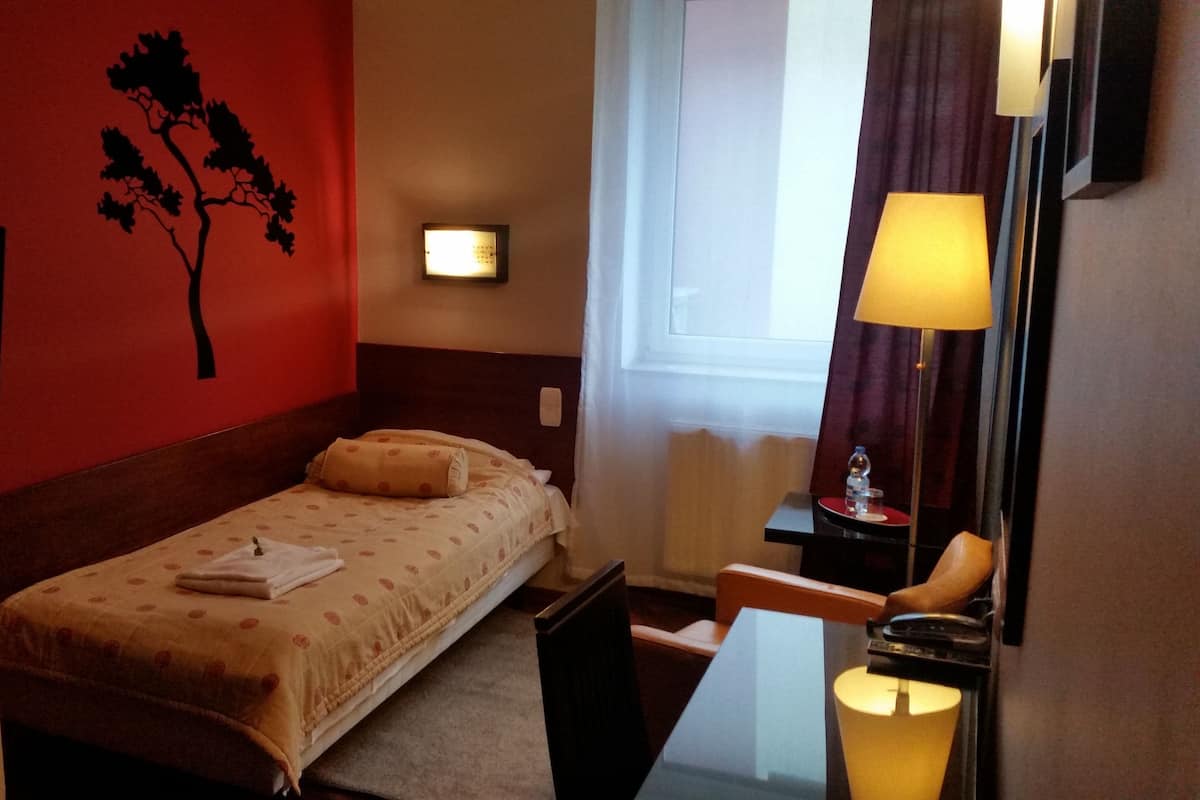 Hotellihuone, jossa on yksi sänky, yöpöytä, työpöytä tuolilla, lamppu ja ikkuna, jossa on verhot.
