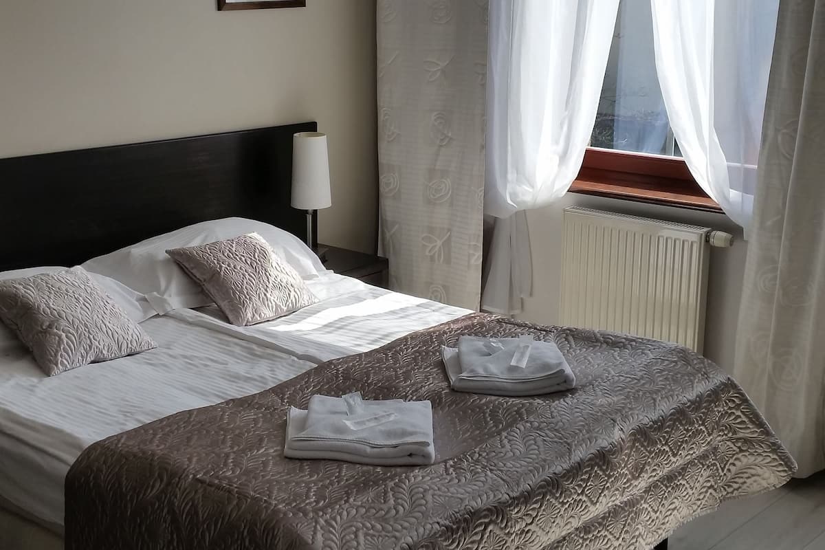 Hotellihuone, jossa on suuri sänky, yöpöytä lampun kanssa, seinällä oleva kehystetty taulu ja läpikuultavat verhot ikkunan edessä.