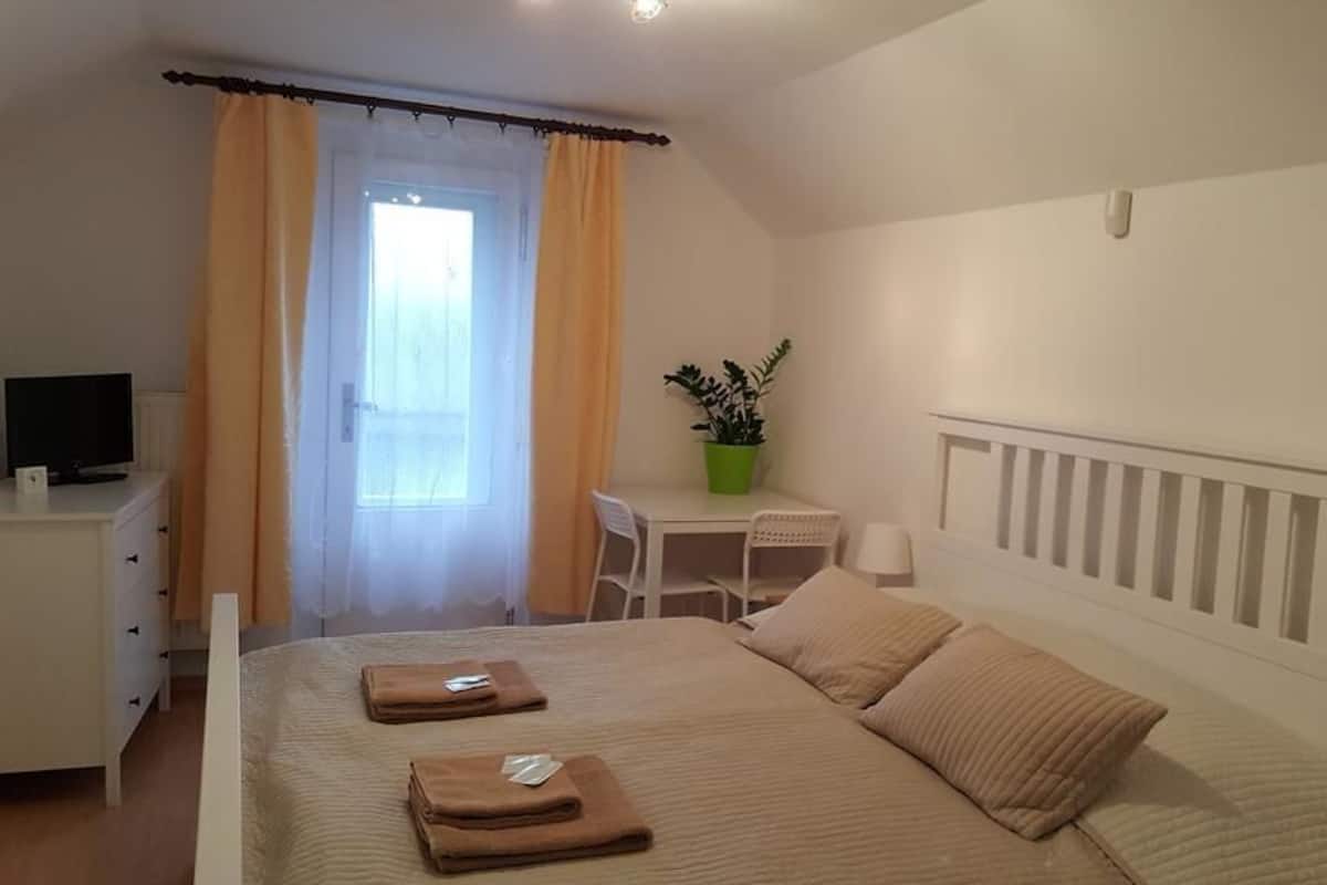 A bedroom with a bed, a desk, a chair, a television, a window with curtains, and a potted plant.