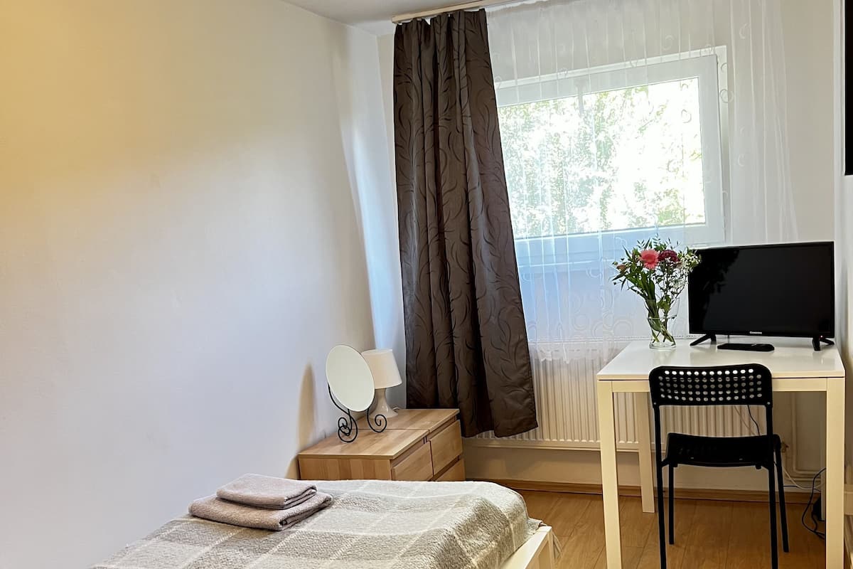 A bedroom with a bed, a desk with a chair, a TV, a lamp, and a window with curtains.