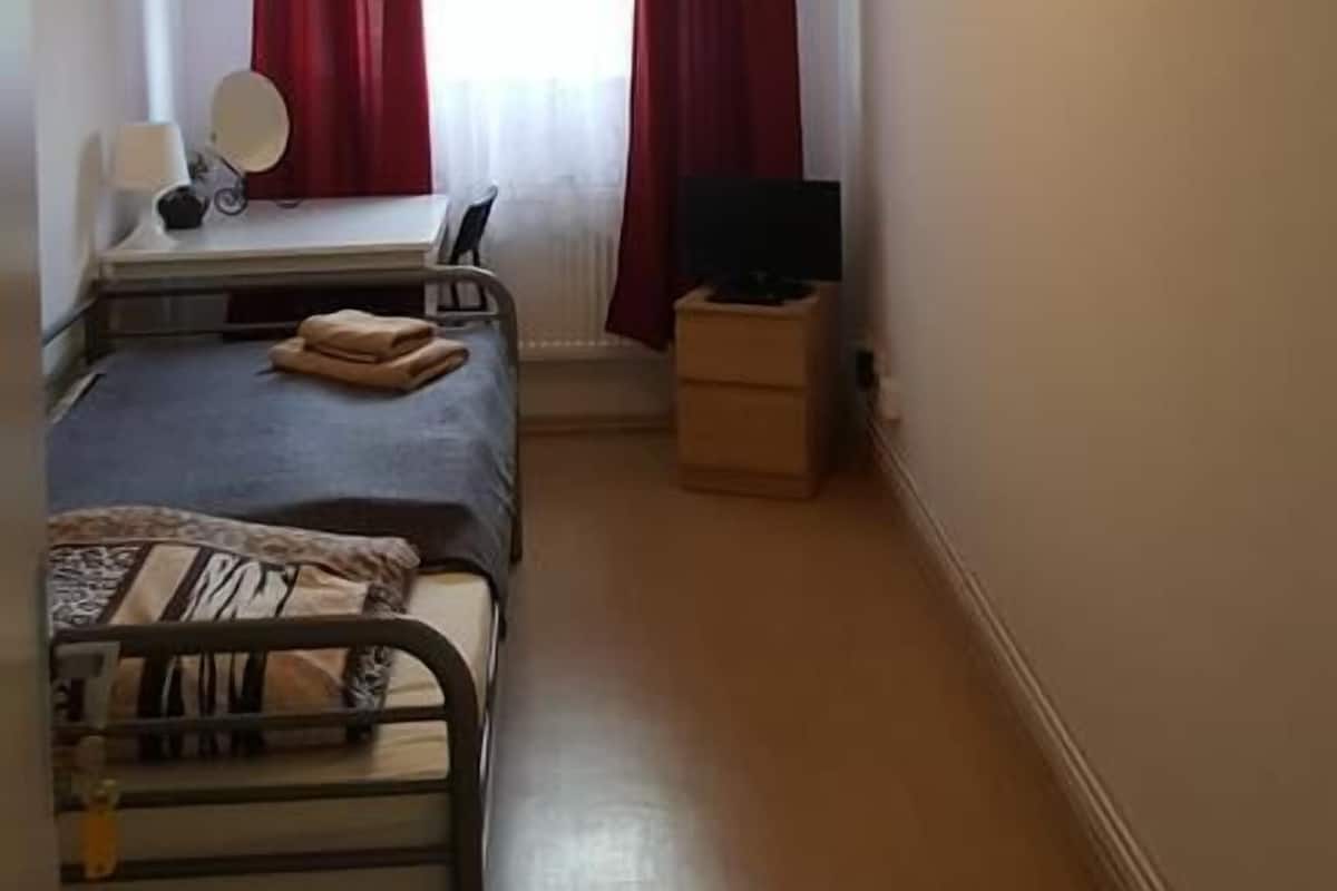 A room with two beds, a desk with a chair, a mirror, and a small table with a lamp. There is a window with curtains and a ceiling light fixture.