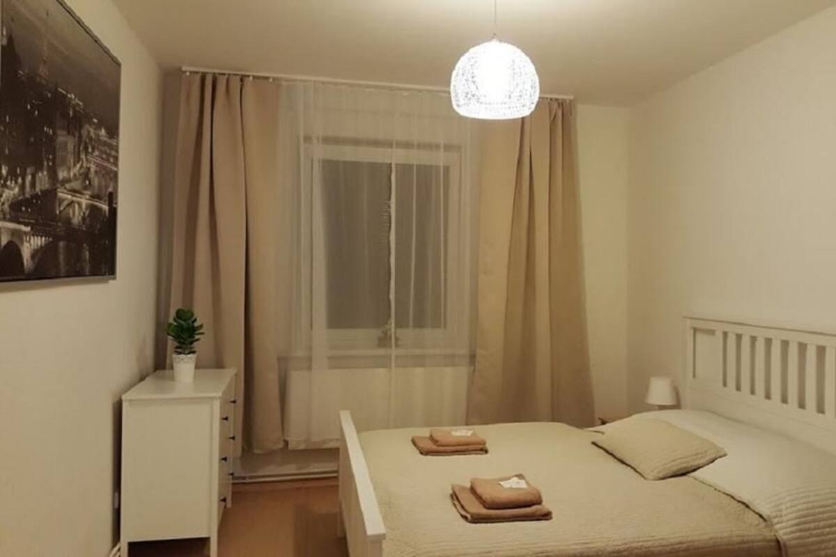 A bedroom with a bed, a bedside table, a hanging light fixture, a window with curtains, and a wall-mounted picture.