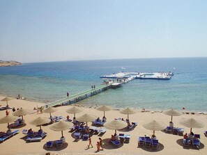 Una playa con tumbonas y sombrillas, un muelle que se adentra en el mar, y barcos amarrados.