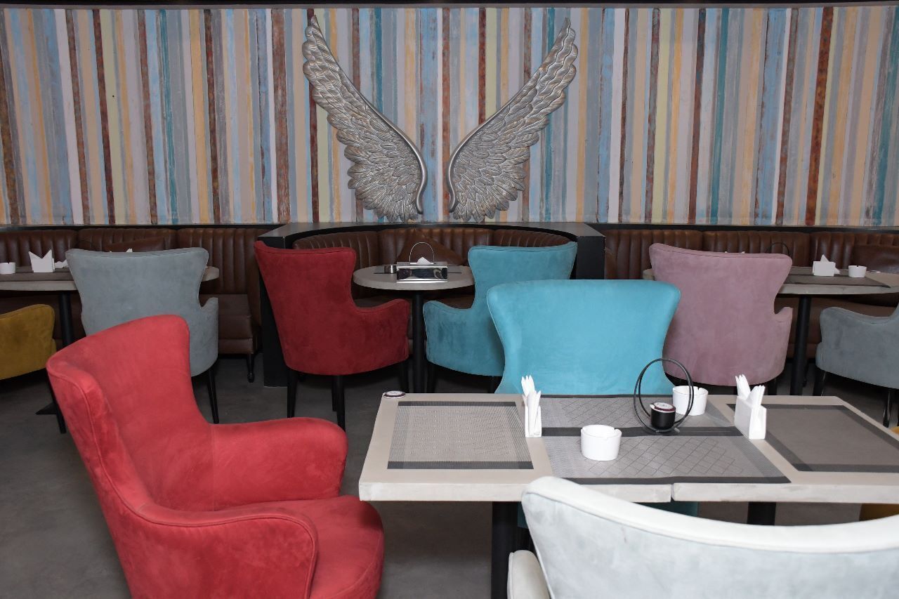 A restaurant interior with cushioned chairs in various colors, a striped wall with a winged sculpture, and tables with napkins and cutlery.