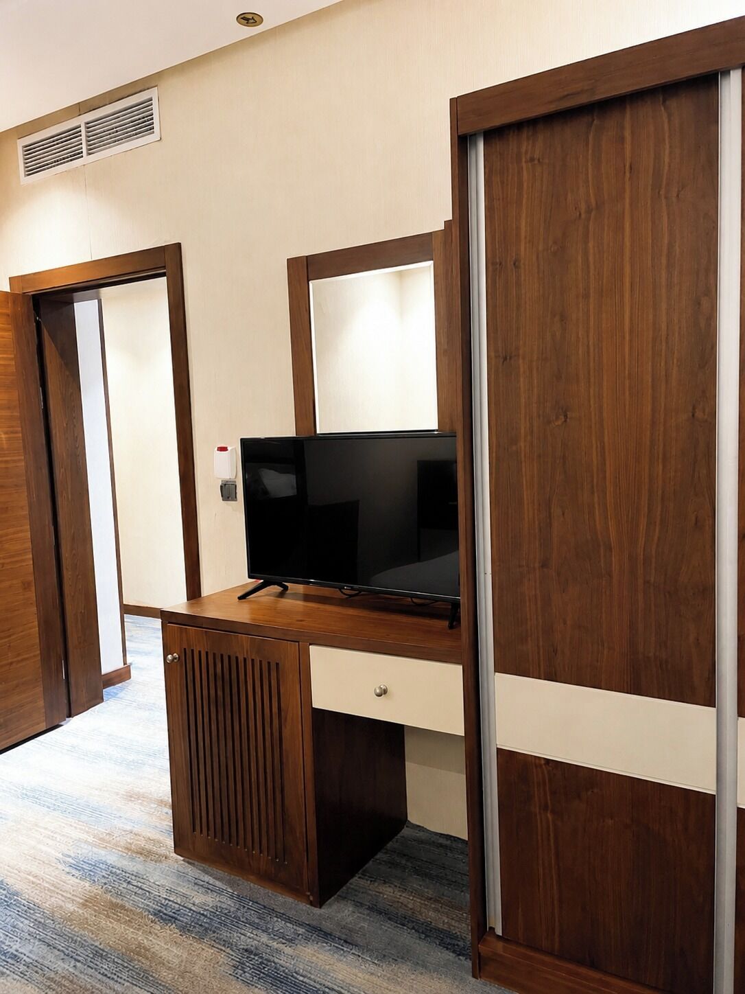 Een kamer met een houten bureau, een televisie, een schuifdeur en een ventilatierooster aan de muur.